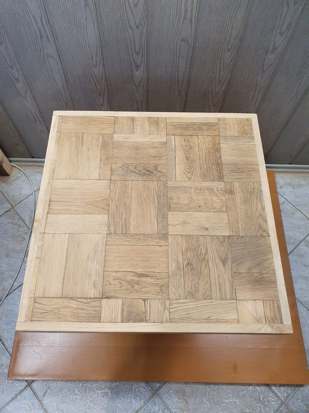 Blat stołu z jasnego drewna ułożonego w kwadraty tworzące wzór parkietu, otoczony ramą z litego drewna, na brązowej podstawie.