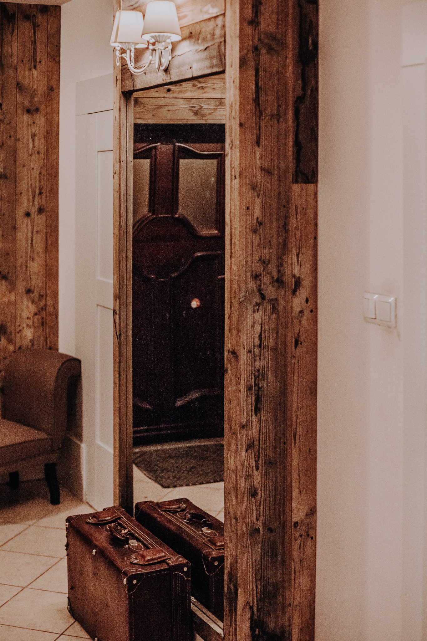 Rustykalne drzwi drewniane z ościeżnicą z grubych belek, widoczne przez odbicie w lustrze, obok dwie stare walizki podróżne.