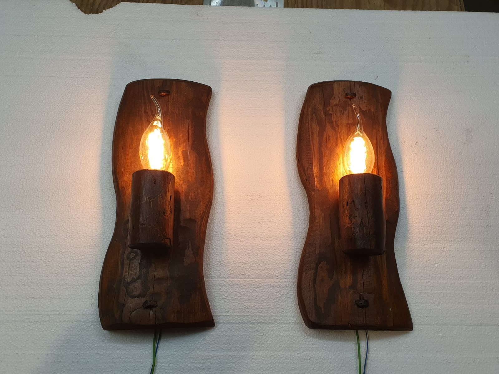 Dwa drewniane kinkiety w stylu rustykalnym z żarówkami w kształcie płomienia, zamontowane na białej ścianie z widocznymi przewodami.
