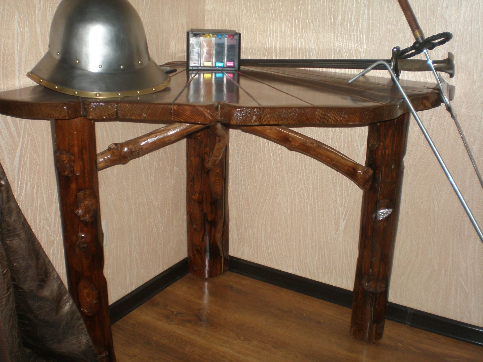 Kanciasty, drewniany stolik z nogami imitującymi gałęzie, na którym leży hełm rycerski i kartridże do drukarki.