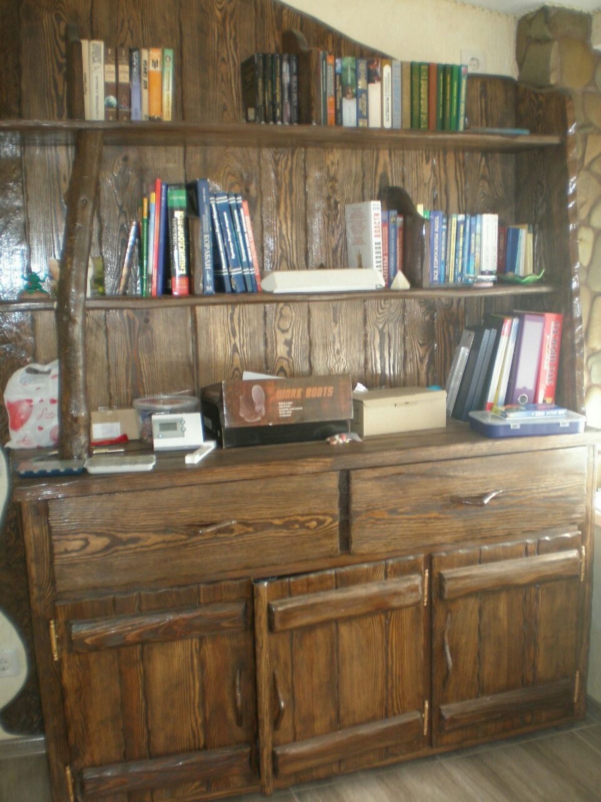 Drewniany regał z półkami i szafkami, wypełniony książkami i drobnymi przedmiotami, stylizowany na rustykalny z widoczną strukturą drewna i gałęzią jako elementem konstrukcyjnym.