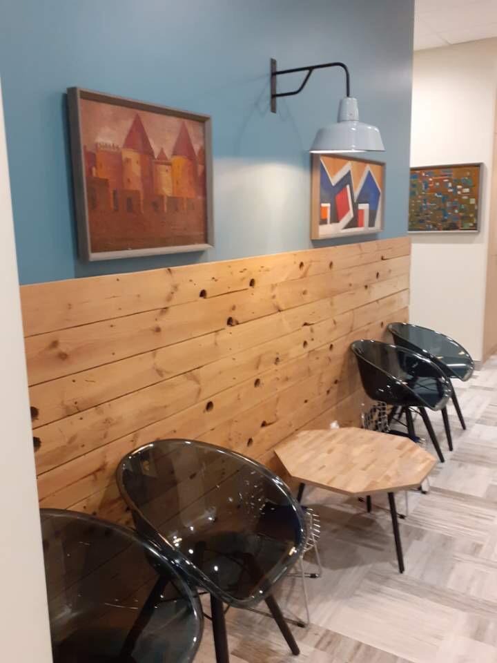 Poczekalnia z drewnianą boazerią, na której wiszą trzy obrazy i lampa w stylu industrialnym. Ustawione są krzesła z przezroczystego tworzywa i mały, drewniany stolik o sześciokątnym blacie.