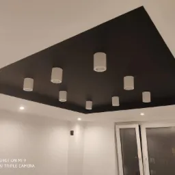 Nowoczesny sufit z czarnym, kwadratowym panelem i rzędem białych, cylindrycznych lamp punktowych, widok z dołu na białe ściany i okno balkonowe.