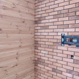 Połączenie drewnianej boazerii i ceglanej ściany z zamontowanym uchwytem do telewizora, widoczne przewody w otworze.