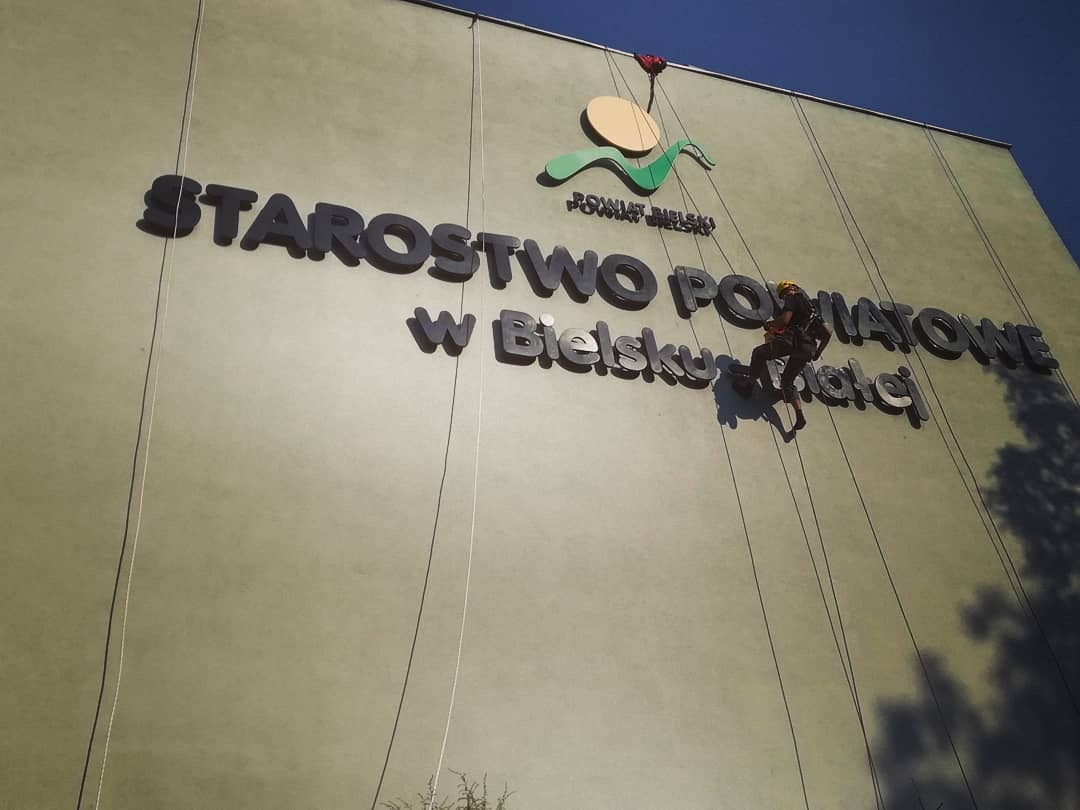 Pracownik na linach zabezpieczających wykonuje prace na elewacji budynku Starostwa Powiatowego w Bielsku-Białej, widoczne duże litery nazwy i logo powiatu.