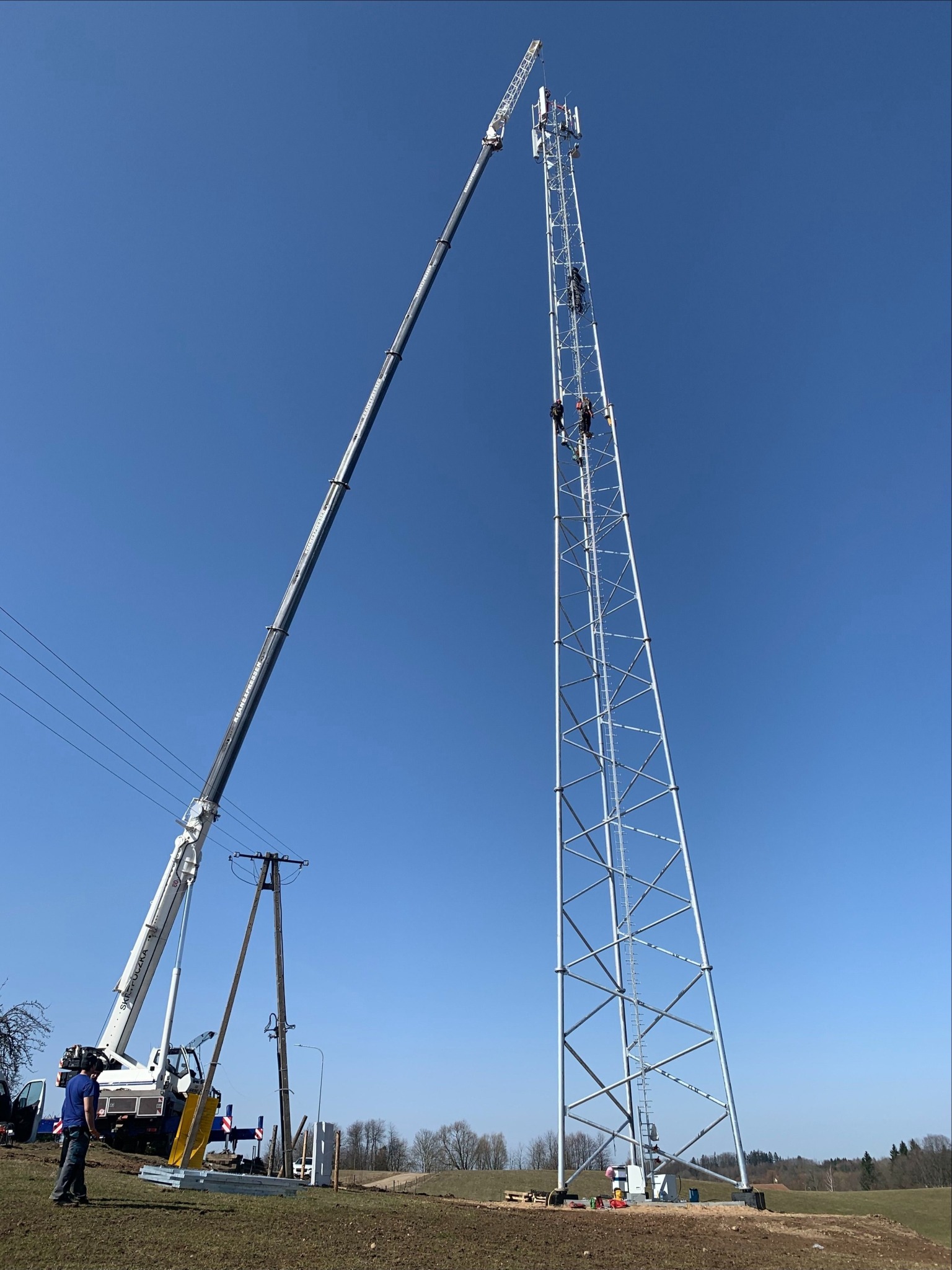Montaż anteny na wieży telekomunikacyjnej z użyciem dźwigu; pracownicy na wysokościach, błękitne niebo w tle.