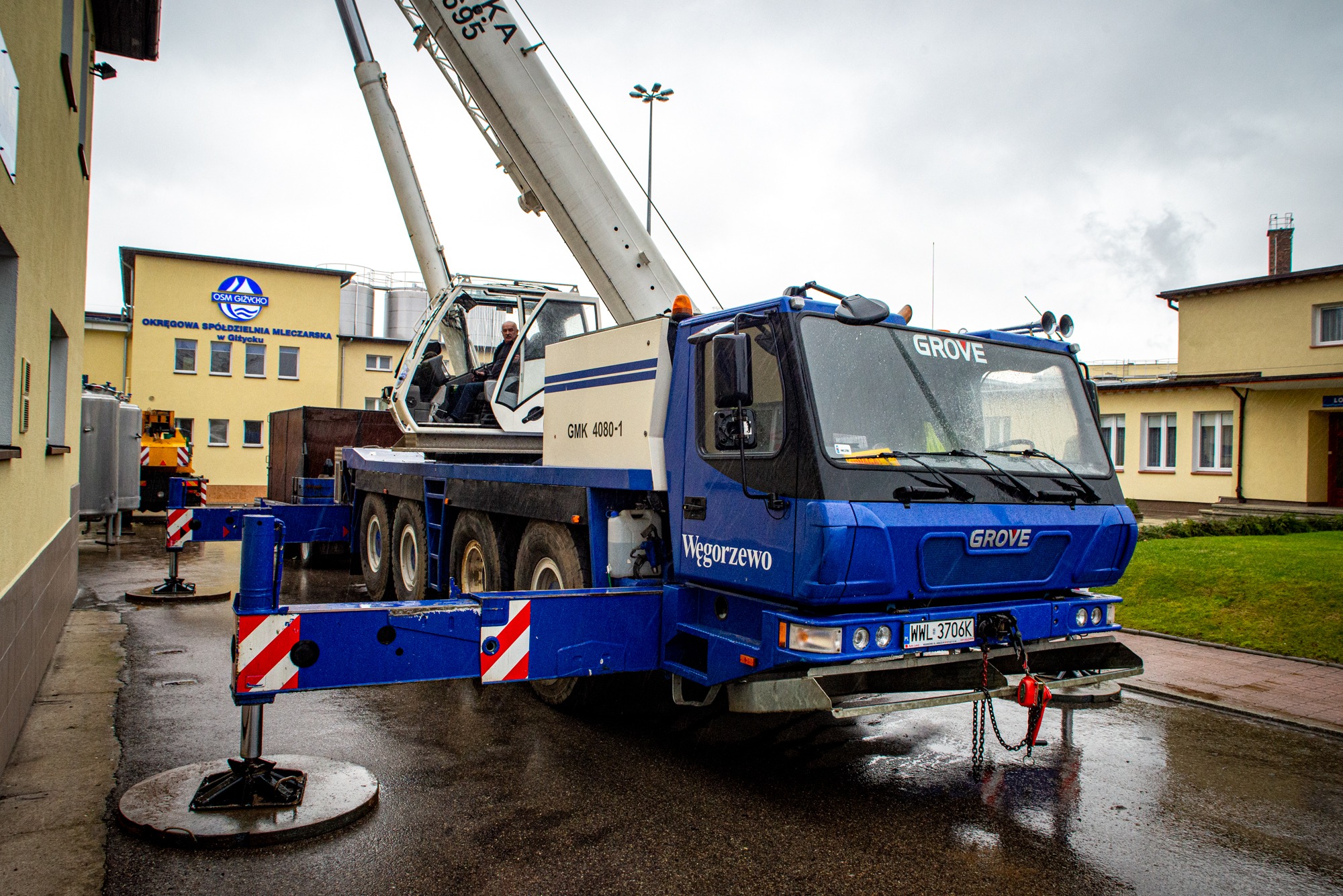 Niebieski dźwig samochodowy Grove GMK 4080-1 z wysuniętym ramieniem i podporami, zaparkowany na mokrym placu przed budynkiem Okręgowej Spółdzielni Mleczarskiej w Giżycku, widoczny operator w kabinie.