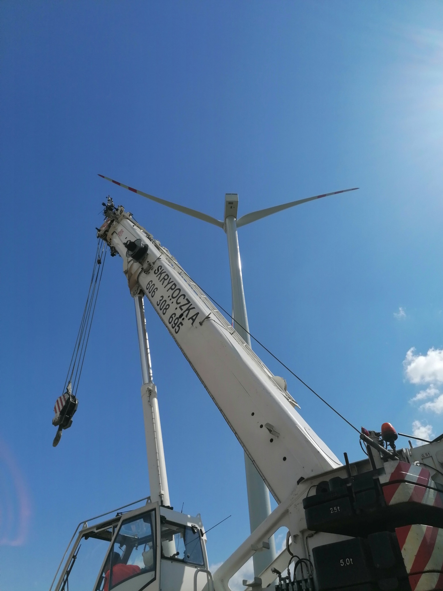 Biały dźwig teleskopowy z wysięgnikiem na tle błękitnego nieba i turbiny wiatrowej w budowie.