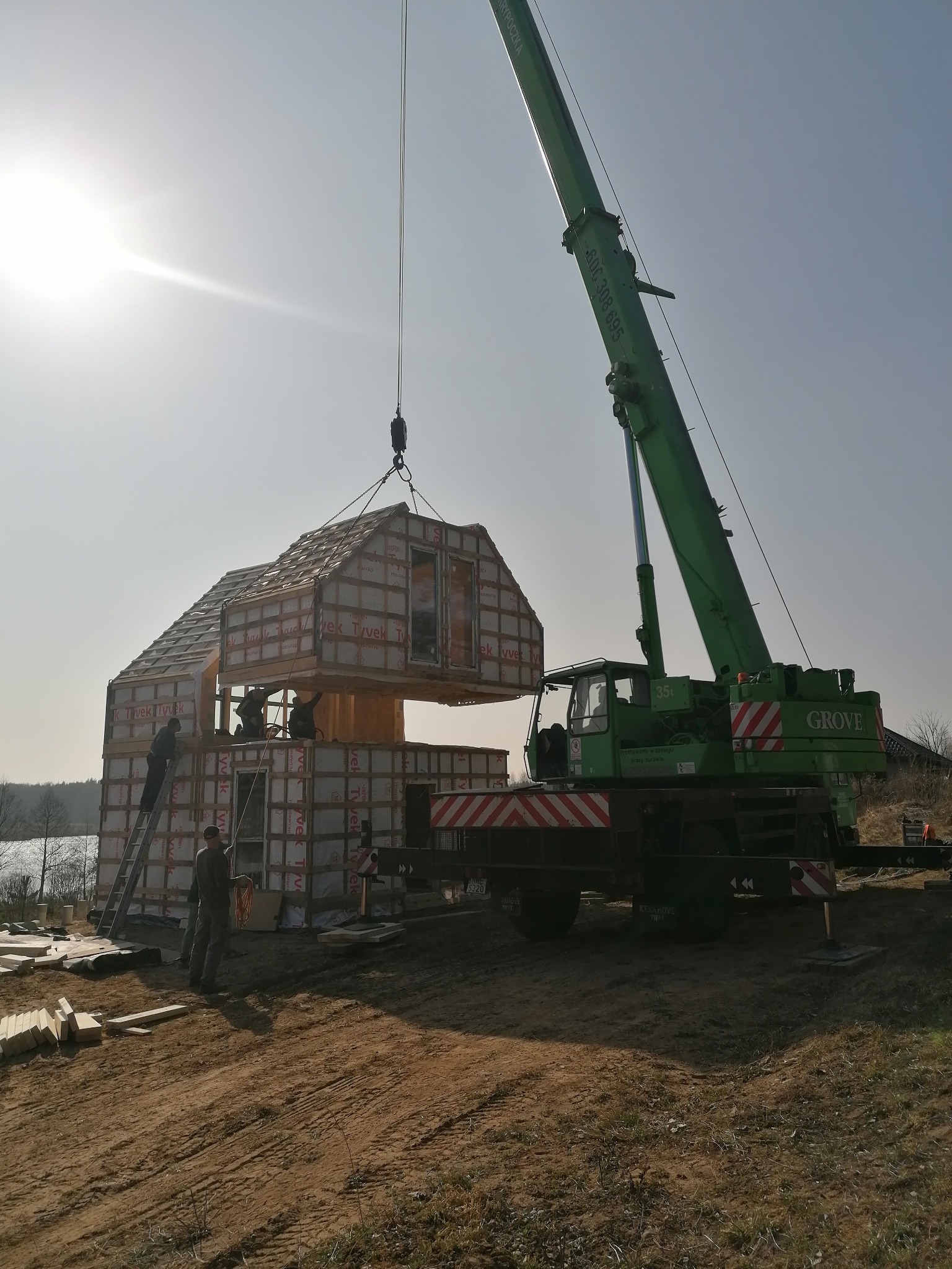 Montaż modułowego domu z użyciem dźwigu Grove 35t, pracownicy na drabinie i wewnątrz konstrukcji, elementy domu owinięte folią Tyvek, widok na jezioro w tle.