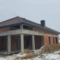 Surowy stan domu jednorodzinnego z czerwonej cegły, z widocznymi betonowymi słupami i częściowo położonym brązowym dachem, na zaśnieżonym terenie.