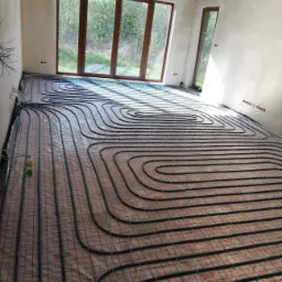 Rozłożona instalacja ogrzewania podłogowego z rurami w kolorze czarnym, widoczne okna i surowe ściany w pomieszczeniu w trakcie budowy.