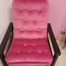 Różowy fotel z weluru z guzikami zdobionymi kryształkami, ciemne drewniane ramy, na tle różowej ściany i jasnej podłogi.