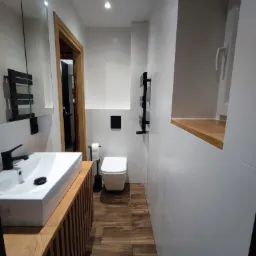 Nowoczesna łazienka z białymi płytkami, drewnianymi akcentami i czarną armaturą, obejmująca umywalkę nablatową, wiszącą toaletę i grzejnik drabinkowy.