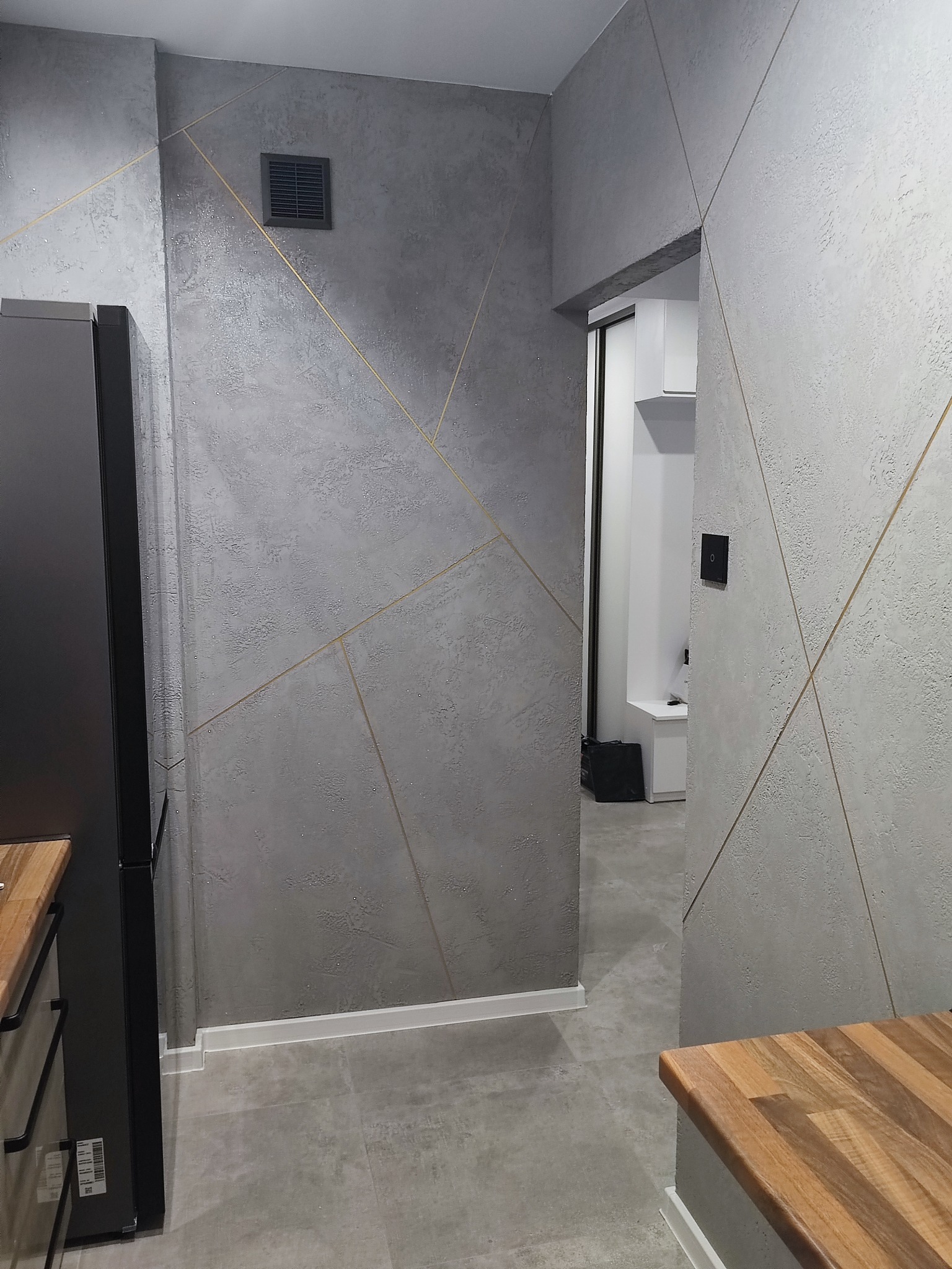 Nowoczesne wnętrze: ściany z dekoracyjnym tynkiem w odcieniach szarości, zdobione złotymi liniami tworzącymi geometryczne wzory; widoczny fragment zabudowy kuchennej i przejście do innego...