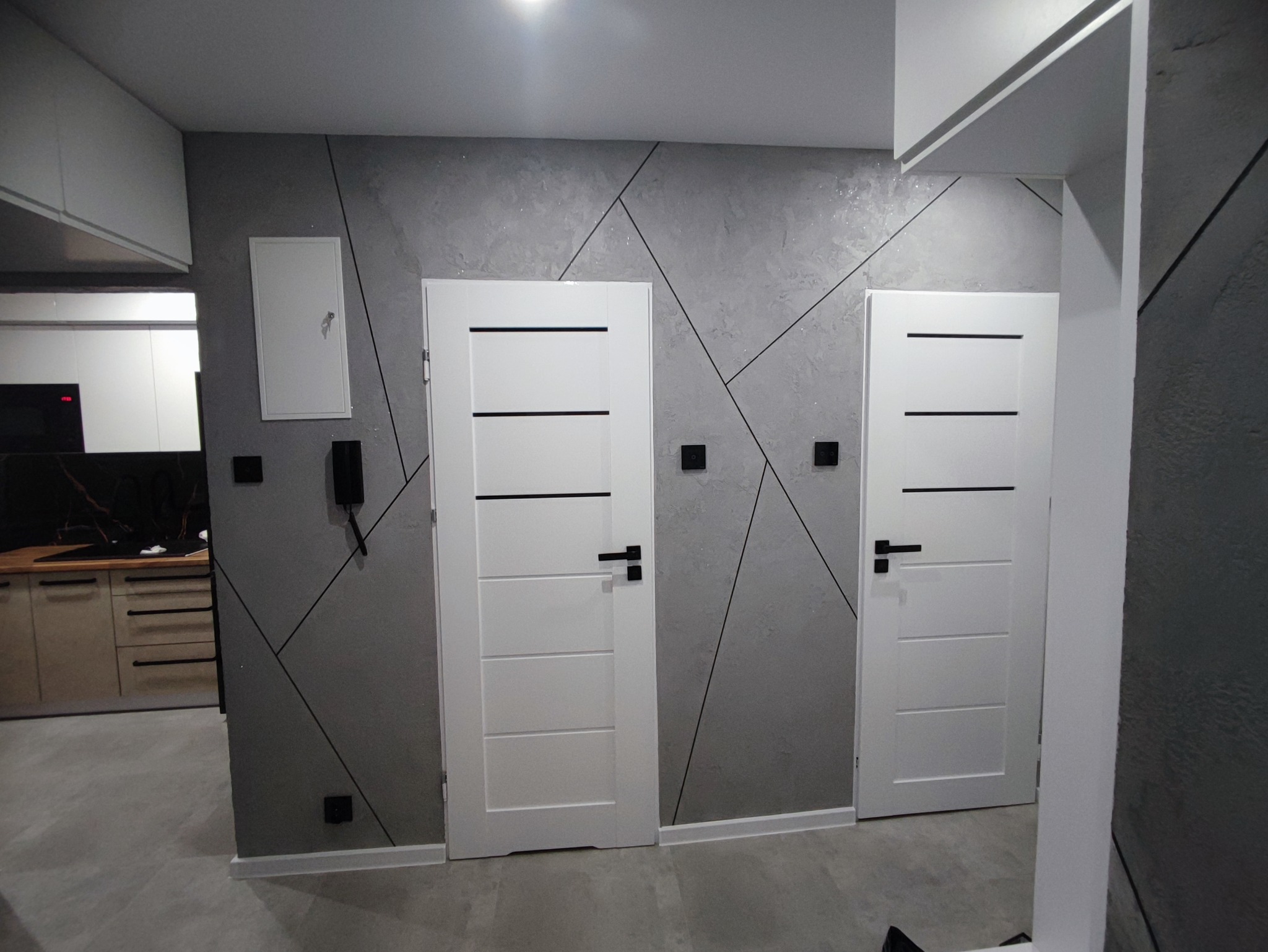 Dwa białe, nowoczesne drzwi wewnętrzne z czarnymi poziomymi paskami, zamontowane w szarej ścianie z geometrycznym wzorem, widok na wykończoną kuchnię w tle.