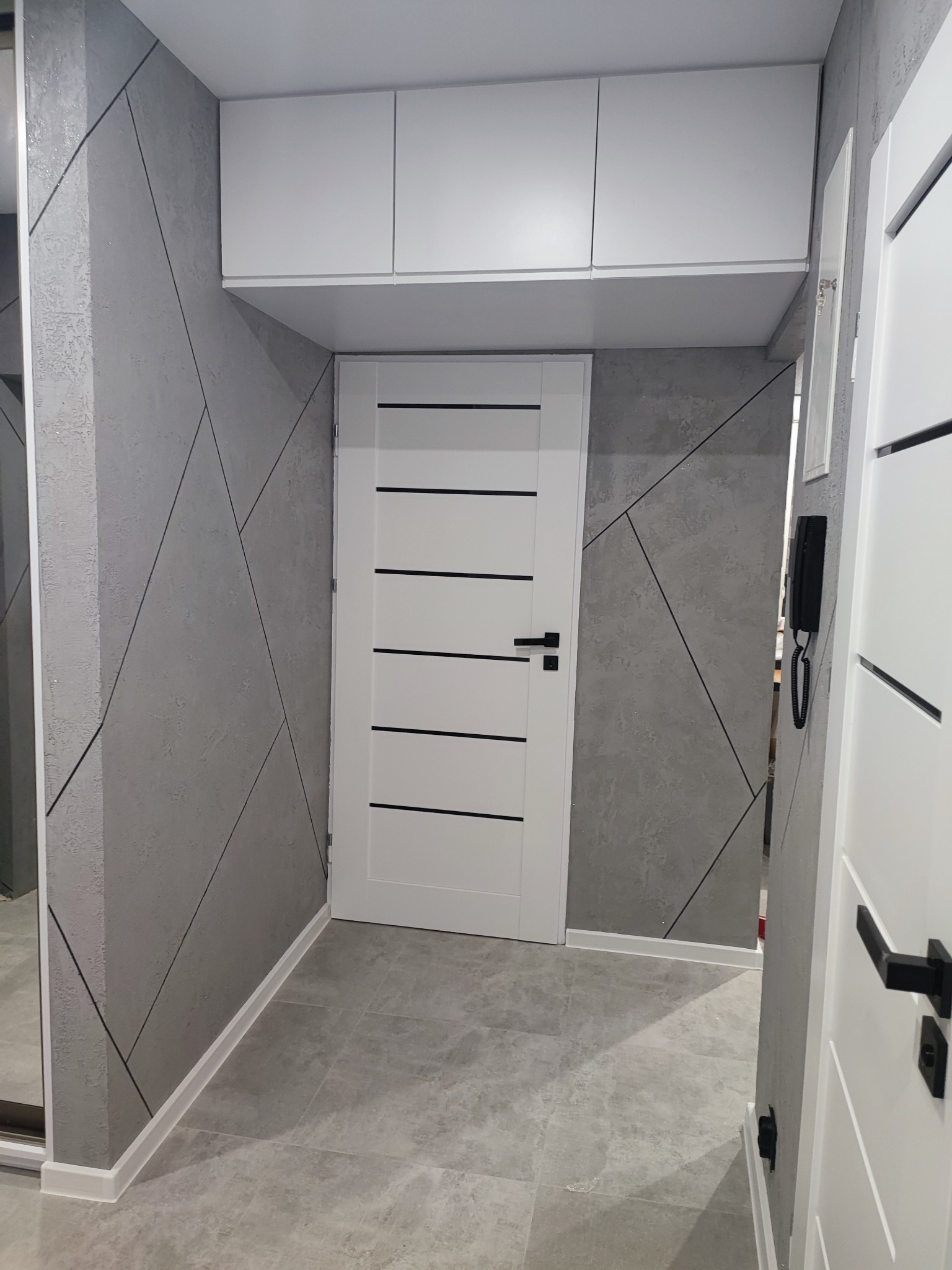 Nowoczesny przedpokój z szarą ścianą o geometrycznym wzorze, białą szafką wiszącą, drzwiami wewnętrznymi z poziomymi czarnymi paskami i szarą podłogą z płytek.