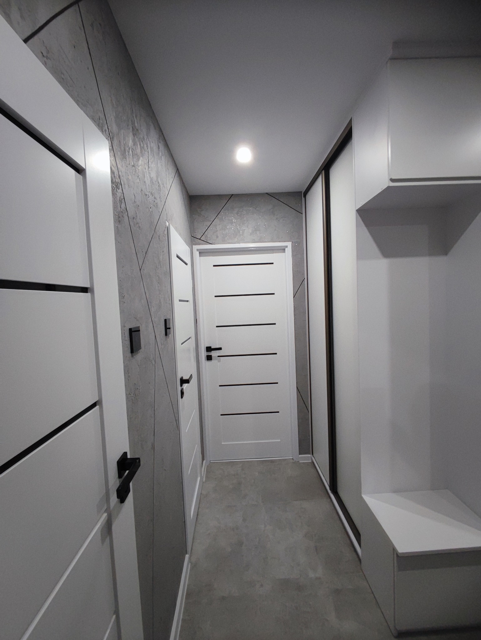 Wnętrze przedpokoju po remoncie: białe drzwi z czarnymi poziomymi paskami, szara ściana z geometrycznym wzorem, szare panele podłogowe, biała szafa przesuwna z siedziskiem.