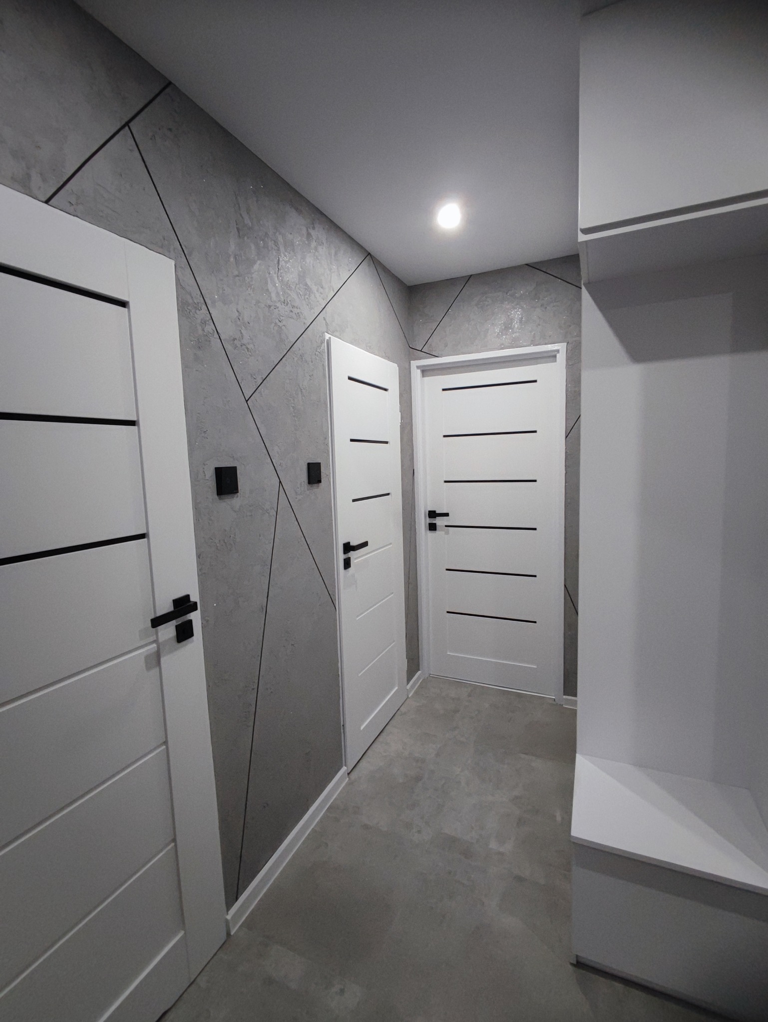 Nowoczesny przedpokój z trzema białymi drzwiami z czarnymi poziomymi paskami i czarnymi klamkami, szara ściana z geometrycznym wzorem, szara podłoga, oświetlenie punktowe