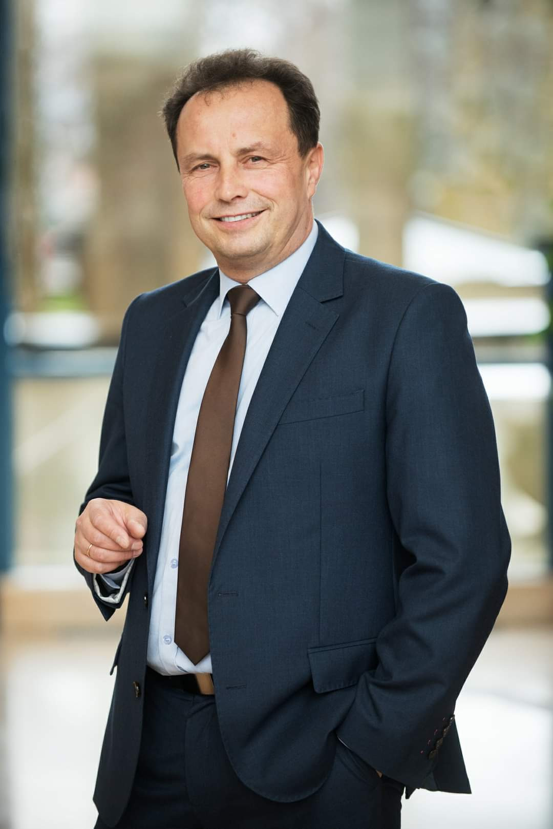 Uśmiechnięty mężczyzna w ciemnym garniturze i brązowym krawacie, pozujący w jasnym wnętrzu, z ręką w kieszeni i okularami w dłoni.