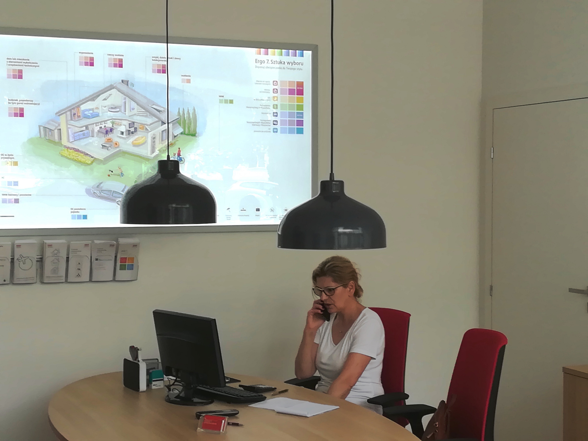 Kobieta w okularach rozmawia przez telefon siedząc przy biurku w biurze, nad nią wiszą dwie czarne lampy, w tle na ścianie wisi grafika przedstawiająca przekrój domu.