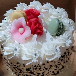 Tort z białym kremem, posypką czekoladową, dekorowany makaronikami i kwiatami z kremu w odcieniach różu i czerwieni, całość umieszczona w kartonowym pudełku.