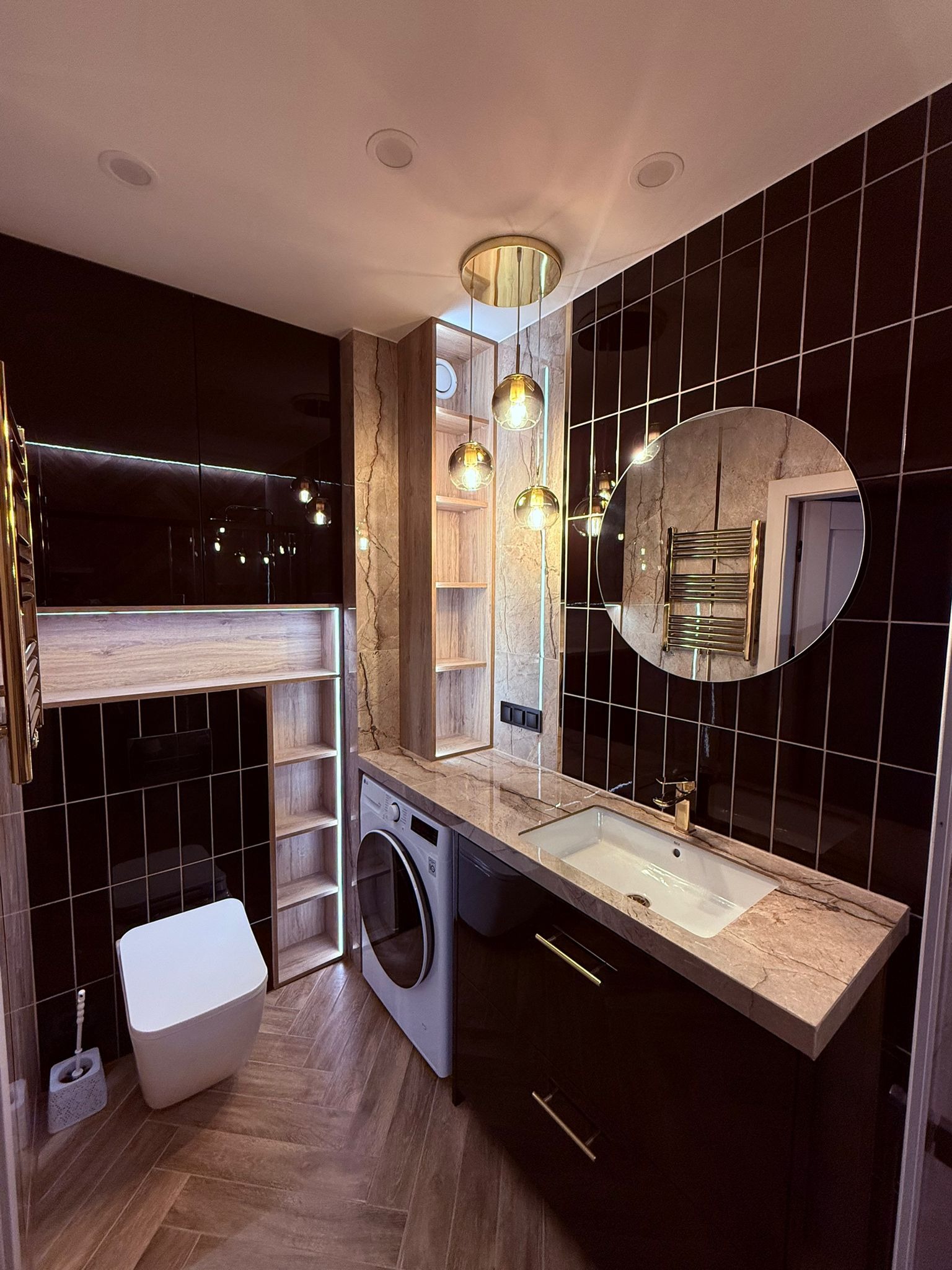 Nowoczesna łazienka z czarnymi płytkami, drewnianą podłogą i marmurowym blatem. Uwagę przykuwa okrągłe lustro i designerska lampa wisząca z trzema szklanymi kloszami.