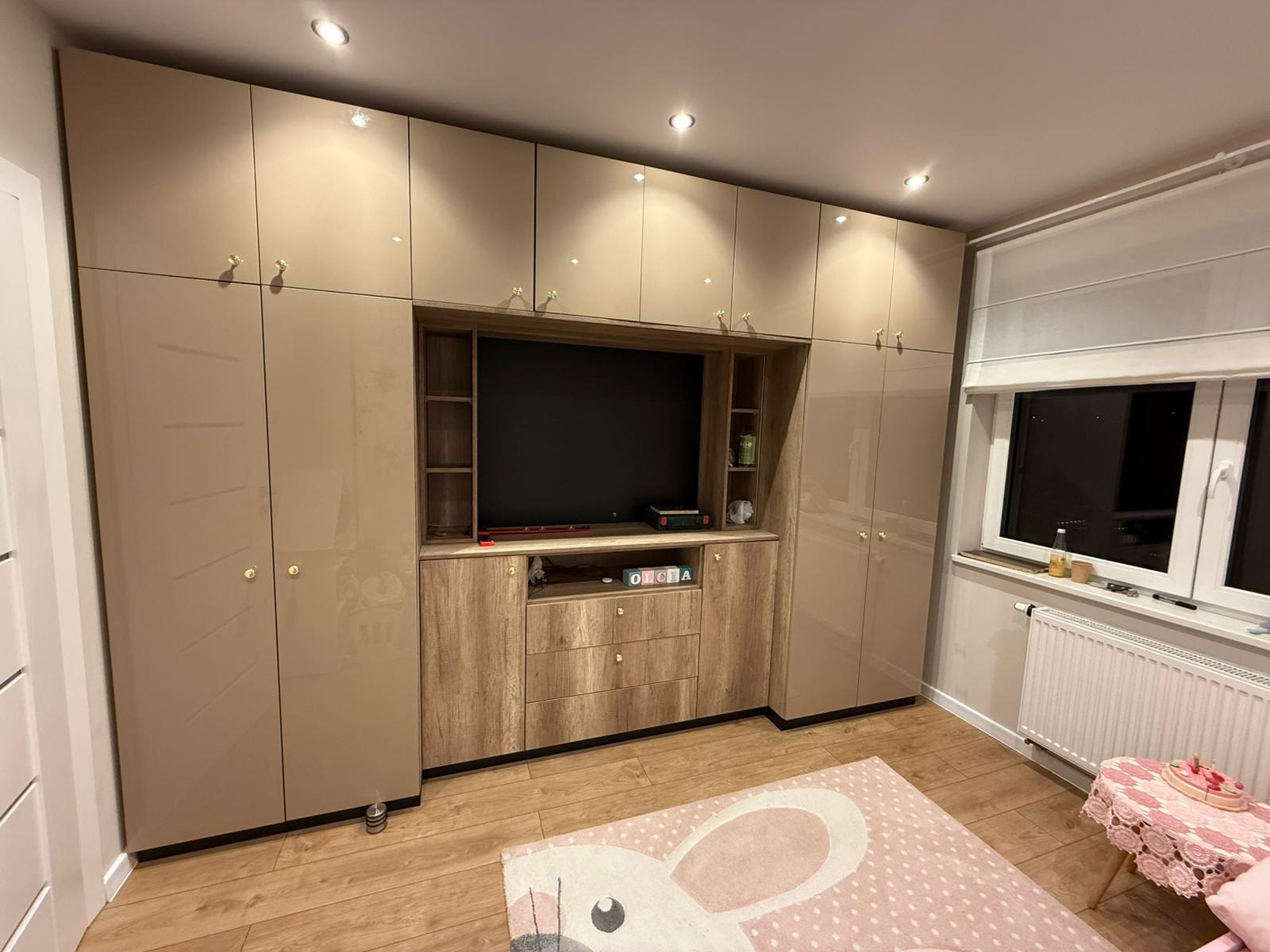 Zabudowa meblowa w pokoju dziecięcym z szafami w beżowym połysku i drewnopodobnym panelem pod telewizor. Widoczne okno z roletą rzymską i drewniana podłoga.
