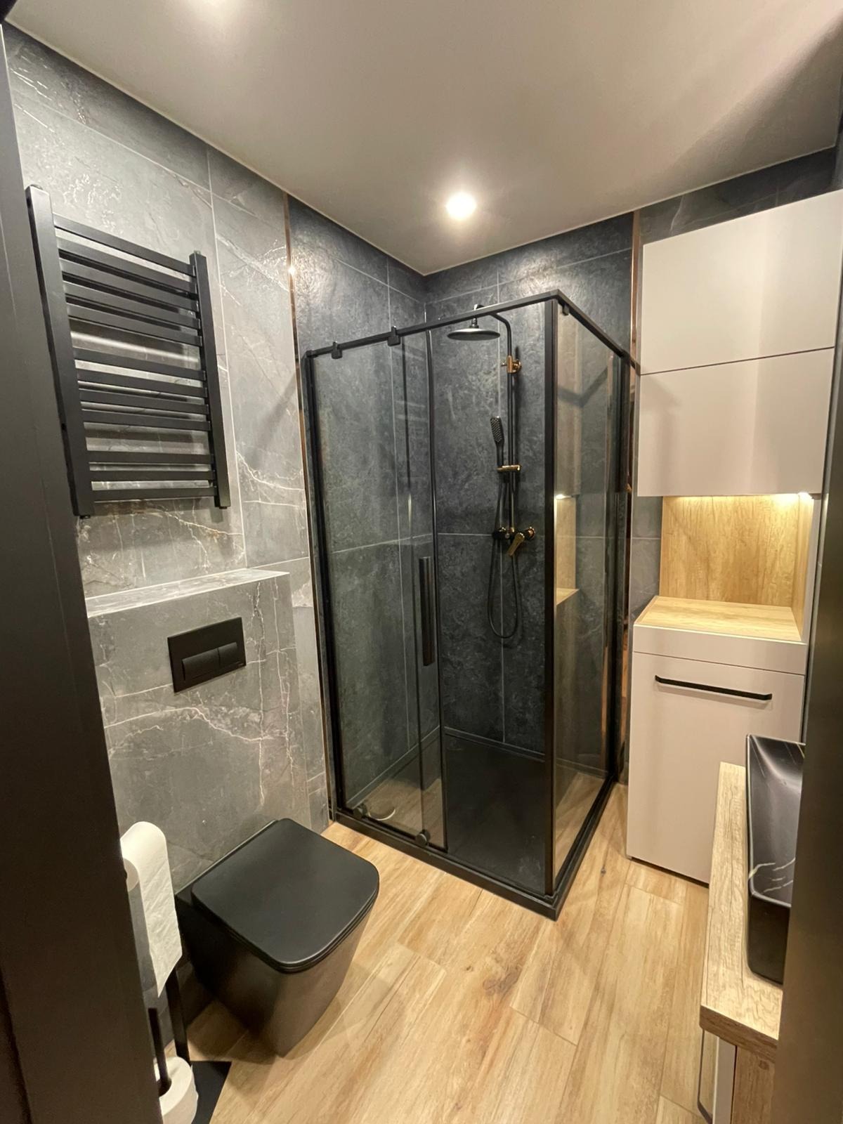 Nowoczesna łazienka z kabiną prysznicową typu walk-in, czarną armaturą i grzejnikiem drabinkowym na tle szarych płytek imitujących marmur. Jasne drewno na podłodze.