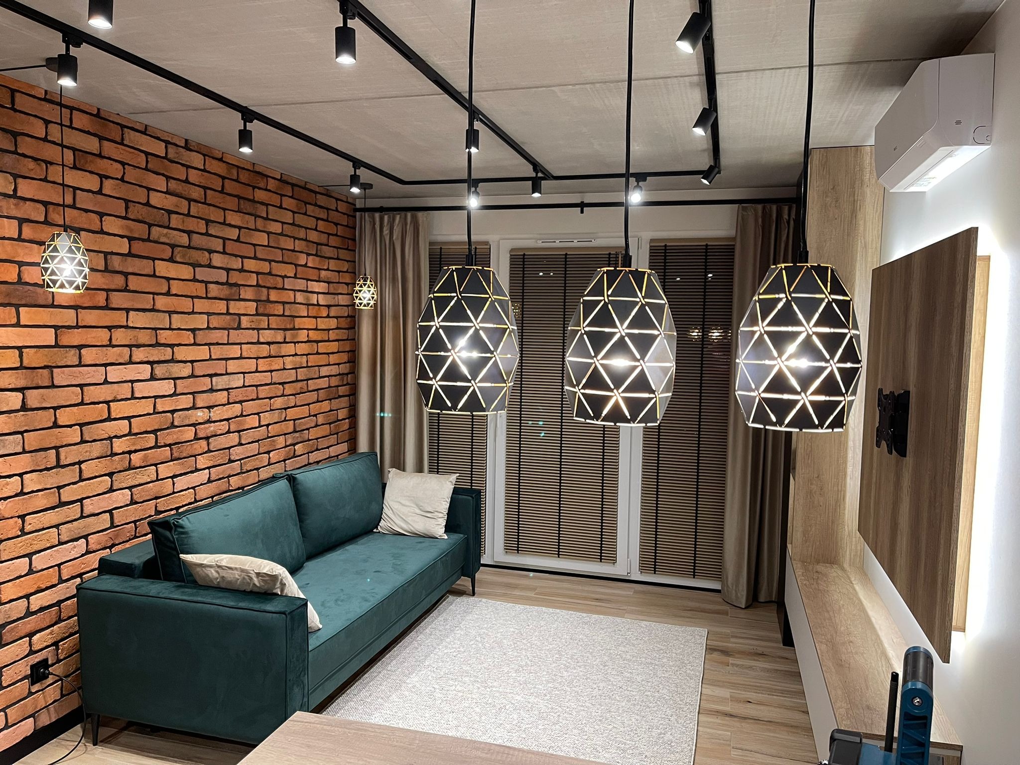 Nowoczesny salon z ceglaną ścianą, zieloną sofą i geometrycznymi lampami wiszącymi. Oświetlenie szynowe na suficie. Widoczne rolety w oknach i drewniane elementy dekoracyjne.