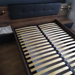 Drewniane łóżko z czarnym zagłówkiem i wbudowanymi lampkami nocnymi, widoczne instrukcje montażu na szafce nocnej.