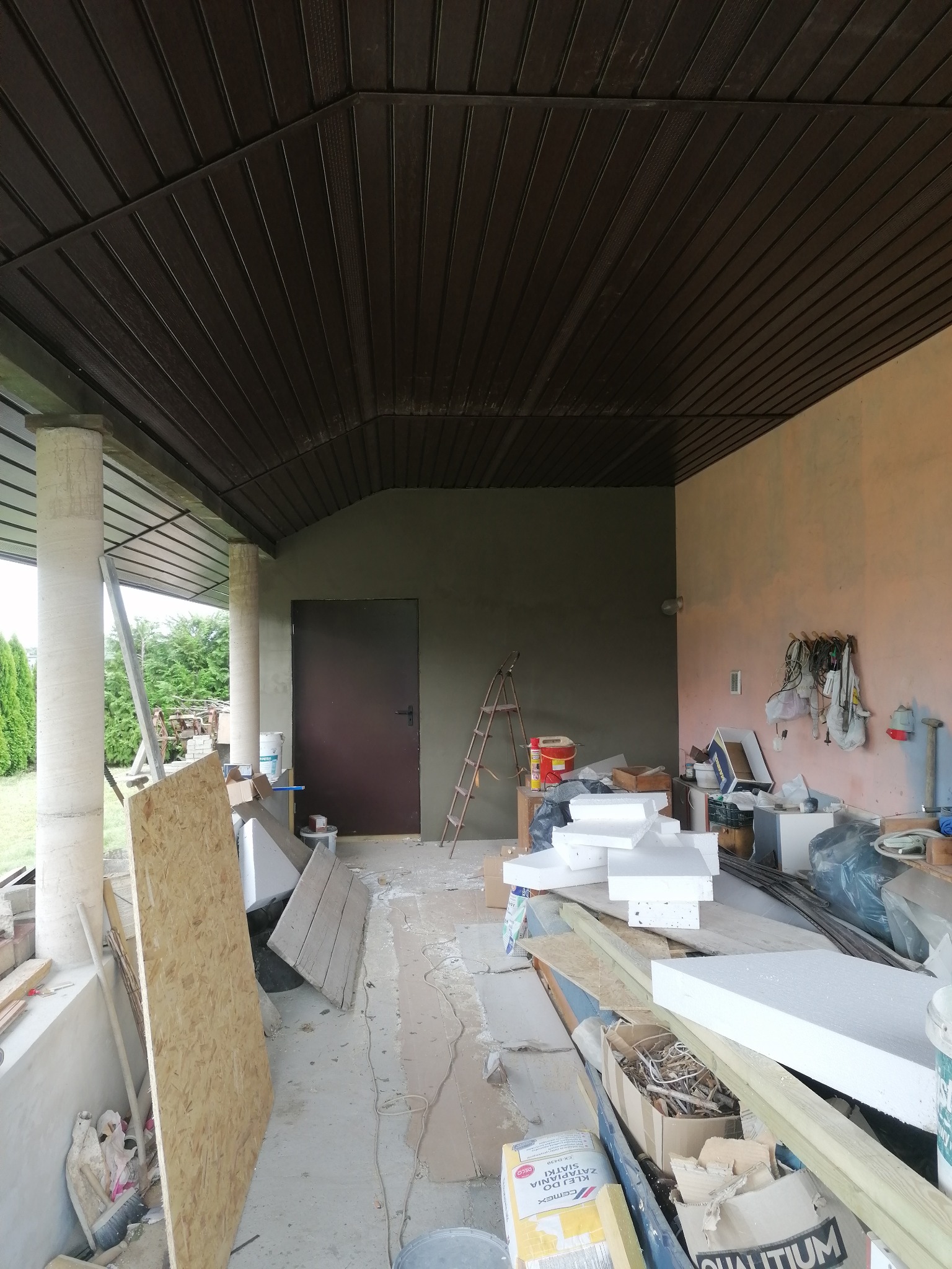 Zadaszony taras w trakcie prac wykończeniowych: sufit z paneli, otynkowana ściana, rozstawiona drabina, materiały budowlane i narzędzia w nieładzie.