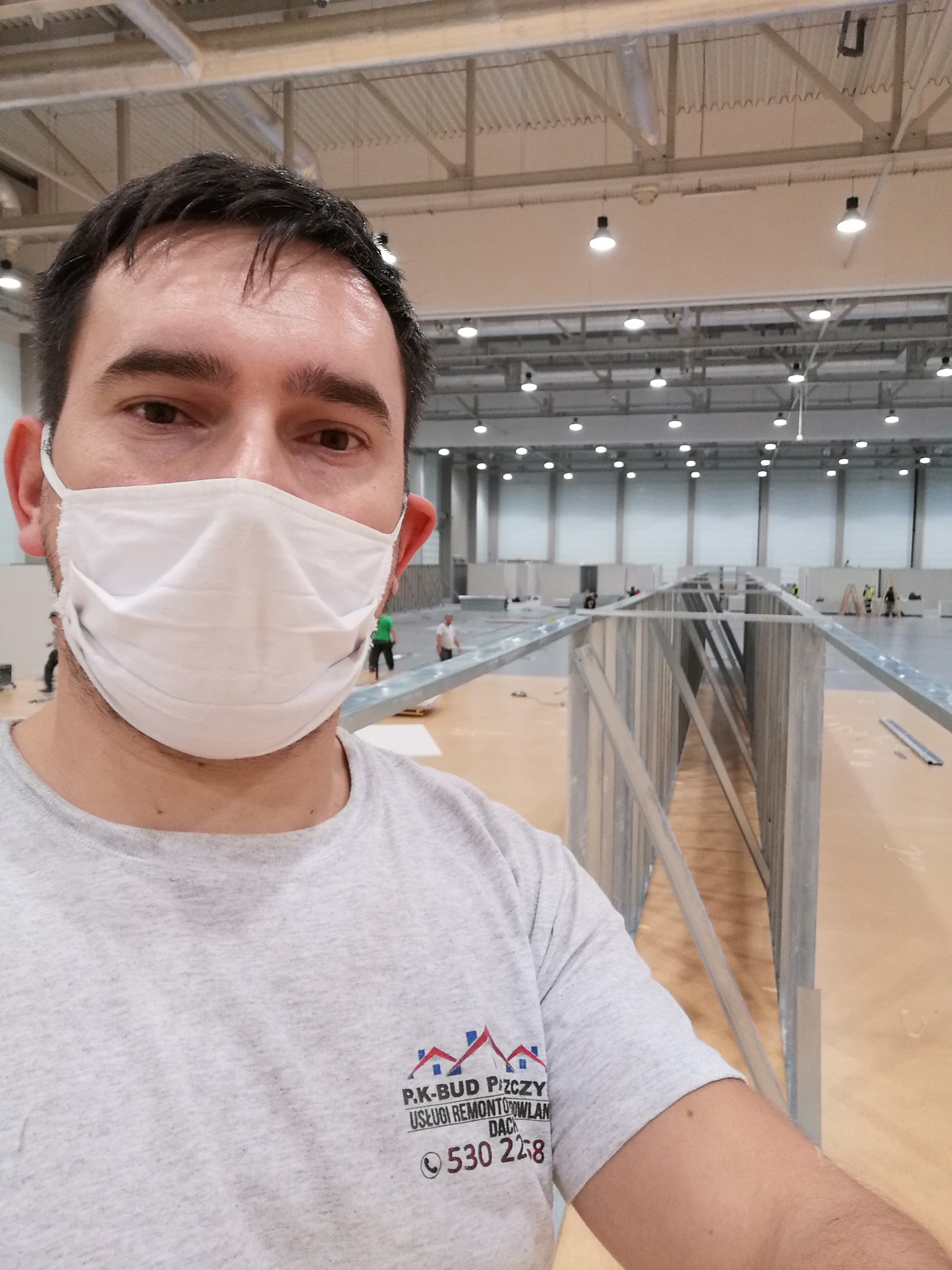 Pracownik w masce ochronnej na tle metalowych konstrukcji ścian działowych w hali, widoczne logo firmy remontowej na koszulce.