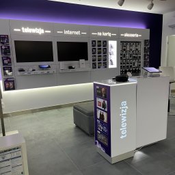 Fach - Wnętrze salonu operatora telekomunikacyjnego z ofertą telewizji, internetu i akcesoriów. Na ścianie ekspozycja telewizorów i akcesoriów. Przy ladzie plakaty z ofertami filmów i seriali.