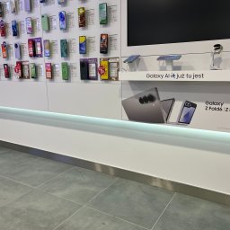 Fach - Ekspozycja smartfonów w sklepie: rząd telefonów z etykietami cenowymi na białej ścianie, poniżej reklama Galaxy AI i Galaxy Z Fold6, szara podłoga z płytek.