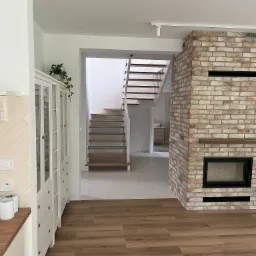 Jasne wnętrze domu z drewnianą podłogą, białymi schodami i ceglanym kominkiem z czarnym wkładem, widok na szafkę z przeszklonymi drzwiami.