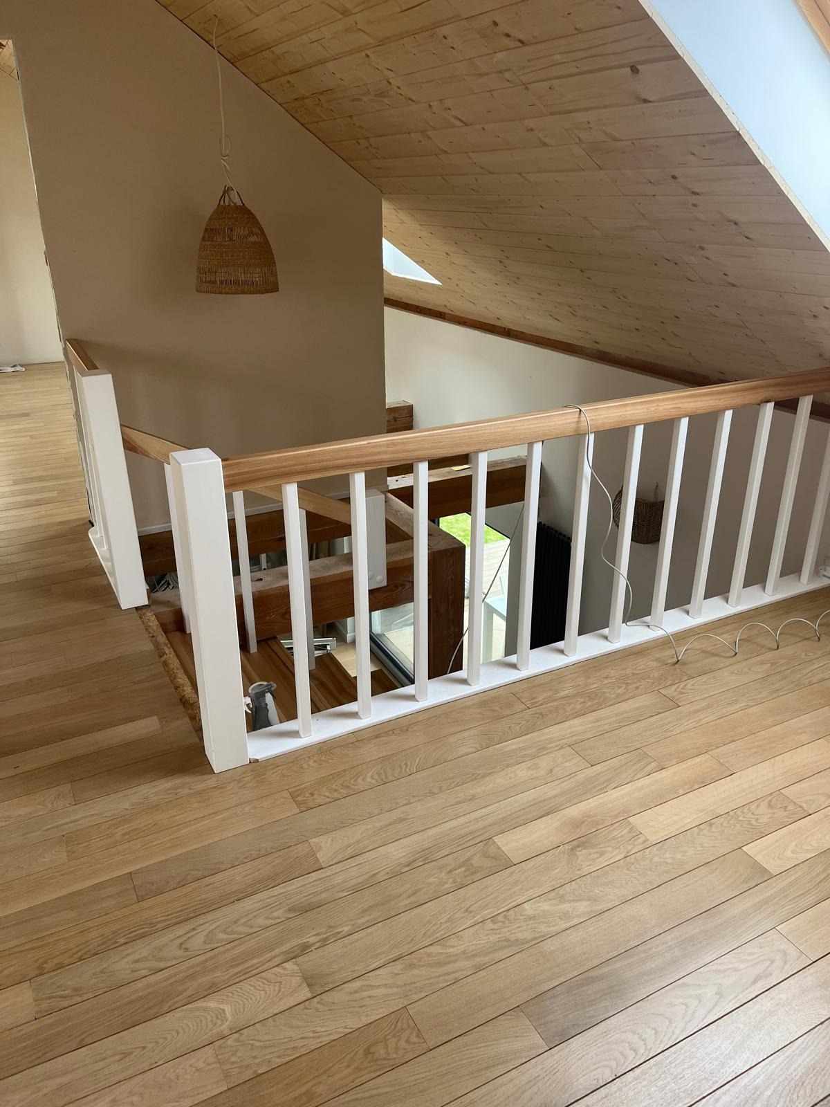 Drewniane schody z białymi balustradami w jasnym wnętrzu poddasza, widoczny fragment okna dachowego i pleciona lampa.
