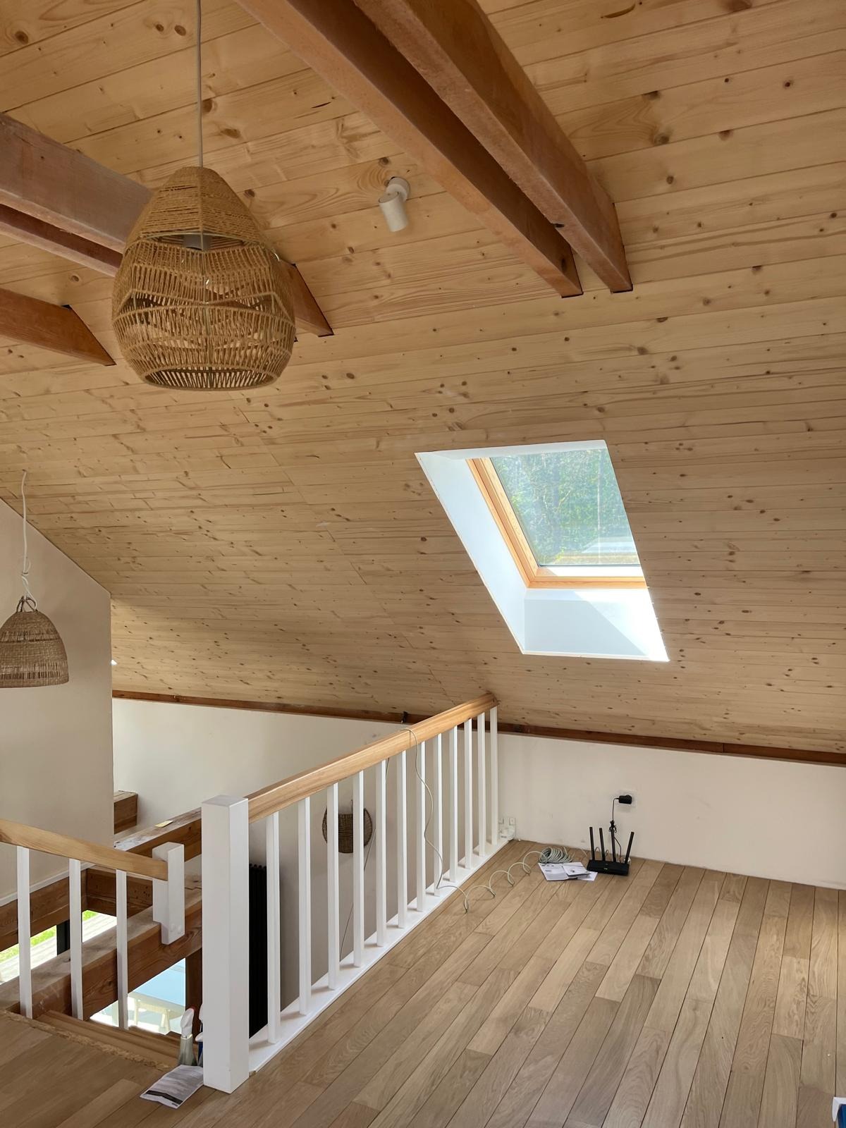 Drewniane wykończenie poddasza z oknem dachowym, widoczne lampy z naturalnego materiału, balustrada schodów i router Wi-Fi na podłodze.