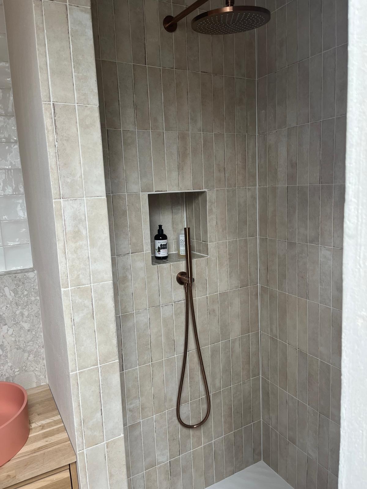 Nowoczesna kabina prysznicowa z beżowymi, pionowo ułożonymi płytkami, wnęką na kosmetyki i miedzianą armaturą.