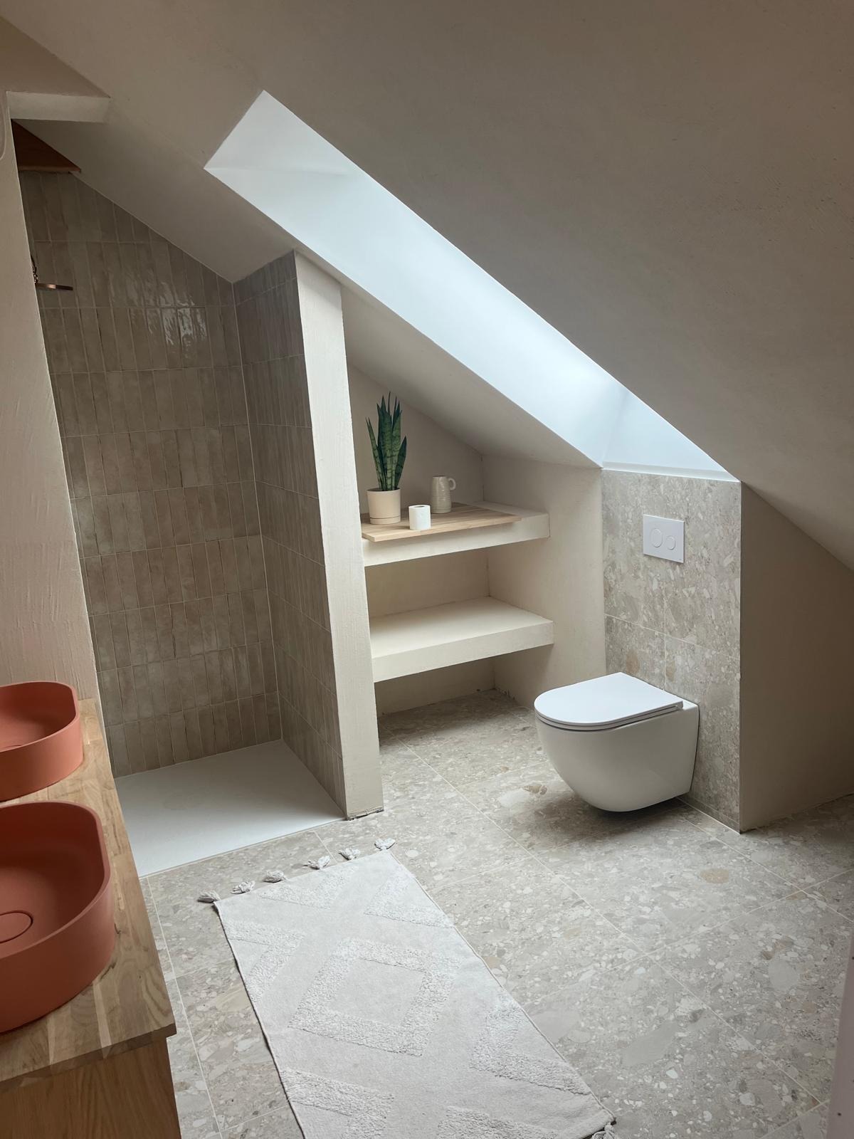 Nowoczesna łazienka na poddaszu z beżowymi płytkami, wiszącą toaletą, kabiną prysznicową bez brodzika, jasnym dywanikiem z frędzlami i rośliną doniczkową na półce pod oknem dachowym.