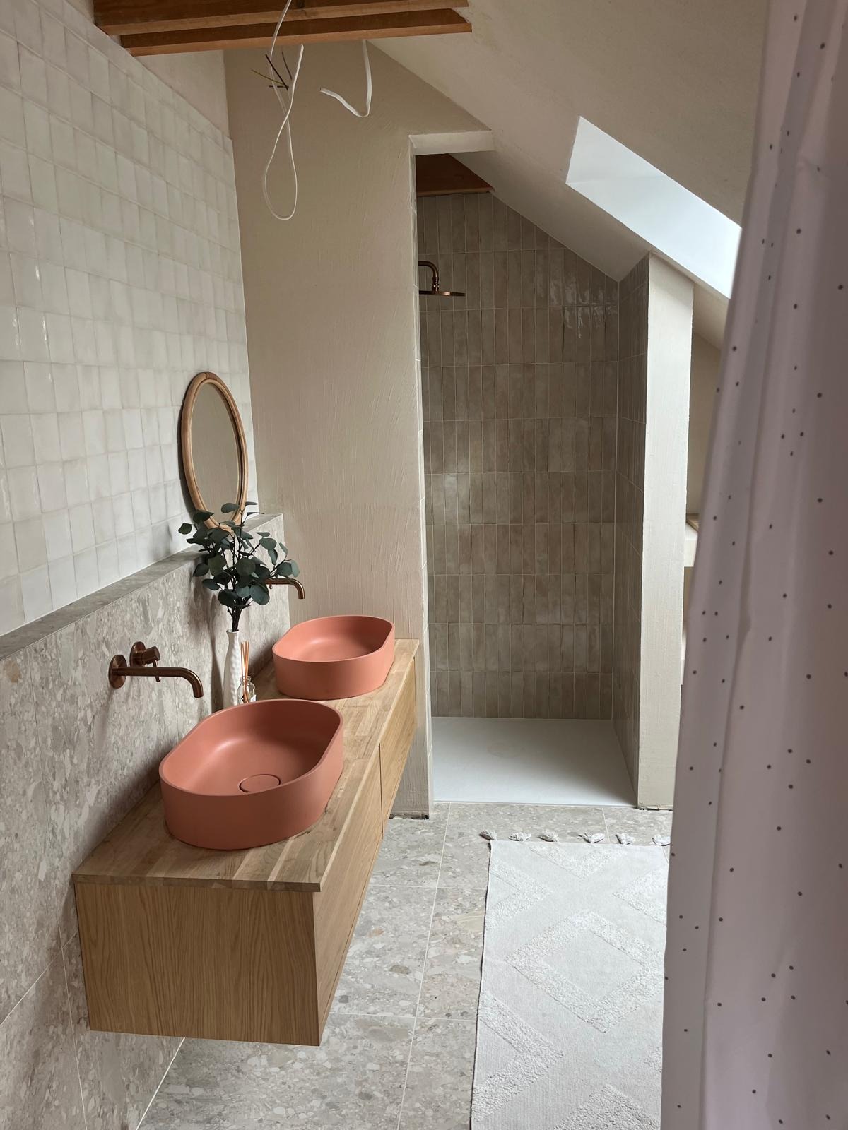 Łazienka na poddaszu z dwiema umywalkami w kolorze terakoty na drewnianej szafce, widoczna kabina prysznicowa z beżowymi płytkami, biały dywanik i fragment zasłony.