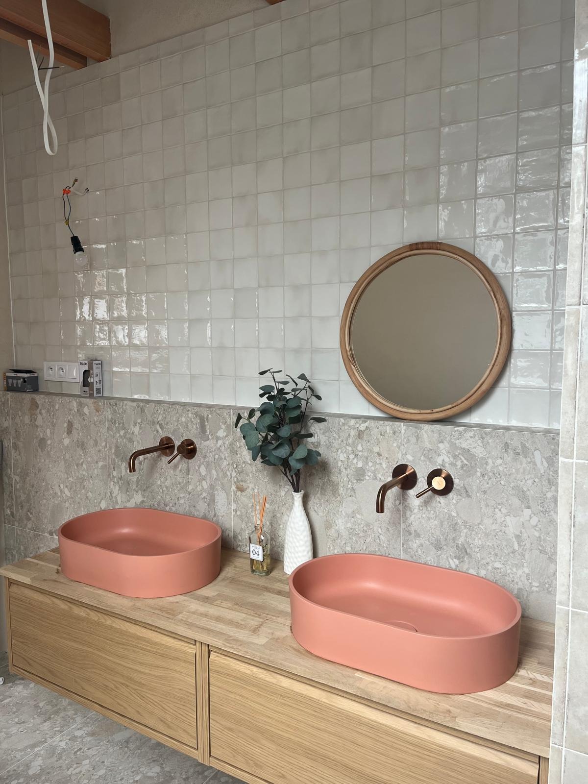 Łazienka w trakcie wykańczania: dwie umywalki w odcieniu brzoskwini na drewnianej szafce, mosiężne krany ścienne, okrągłe lustro w ramie z rattanu, białe kwadratowe płytki na ścianach, widoczne...