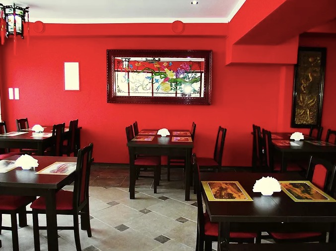 Wnętrze restauracji z czerwonymi ścianami, ciemnymi drewnianymi stołami nakrytymi serwetkami i obrazami w orientalnym stylu.