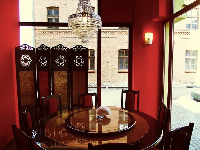 Elegancka sala jadalna z okrągłym stołem, obrotową tacą, krzesłami z czerwonym obiciem, ozdobnym parawanem i kryształowym żyrandolem, widok na ceglany budynek przez okno.