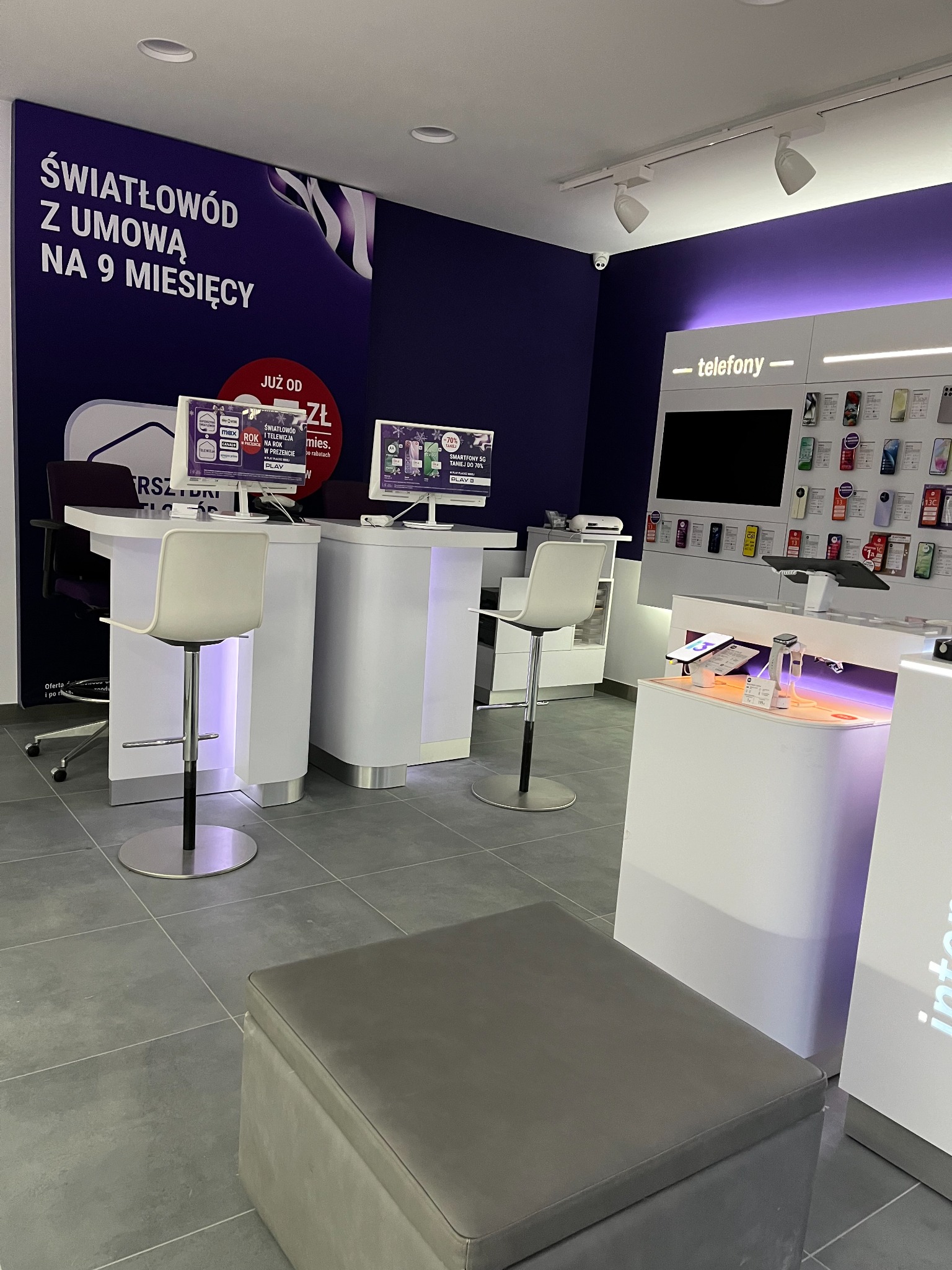Wnętrze punktu sprzedaży operatora telekomunikacyjnego: białe stanowiska obsługi klienta z krzesłami barowymi, ekspozycja telefonów komórkowych na ścianie, fioletowe akcenty kolorystyczne i reklama...