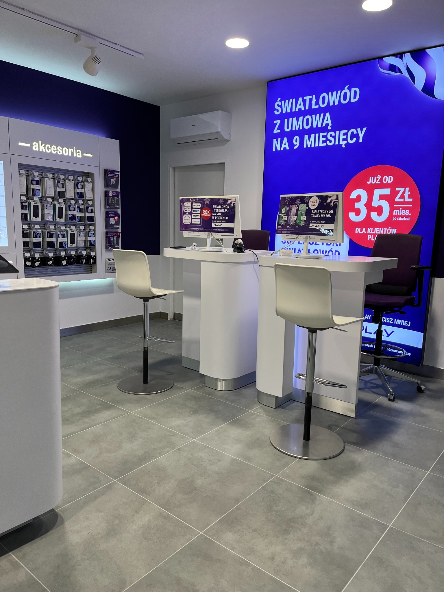 Wnętrze punktu obsługi klienta operatora telekomunikacyjnego z białymi ladami, hokerami i dużym ekranem reklamującym ofertę światłowodu z umową na 9 miesięcy.