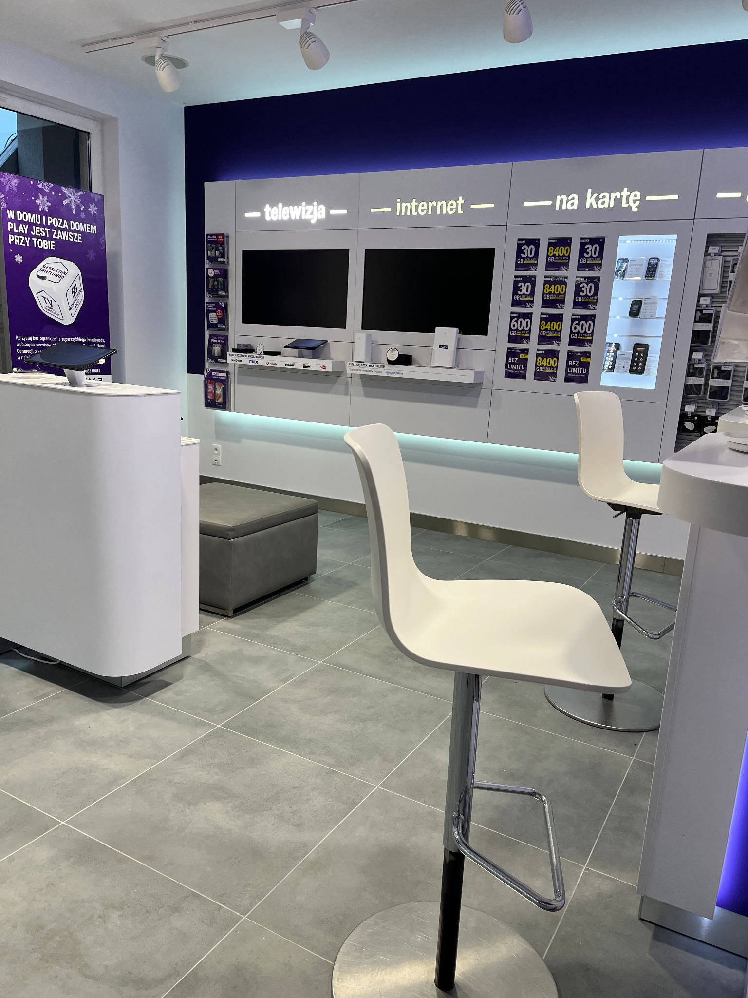 Wnętrze punktu obsługi klienta operatora telekomunikacyjnego z ofertą telewizji, internetu i usług na kartę, widoczne białe lady i krzesła barowe na tle ekspozycji produktów i promocji.