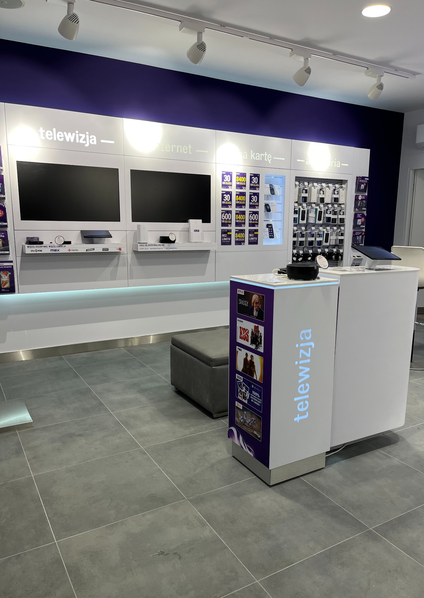 Wnętrze salonu operatora telekomunikacyjnego z ekspozycją telewizorów, dekoderów i akcesoriów telefonicznych podświetlonych ledami, z widocznym napisem 'telewizja' na jednym z mebli ekspozycyjnych.