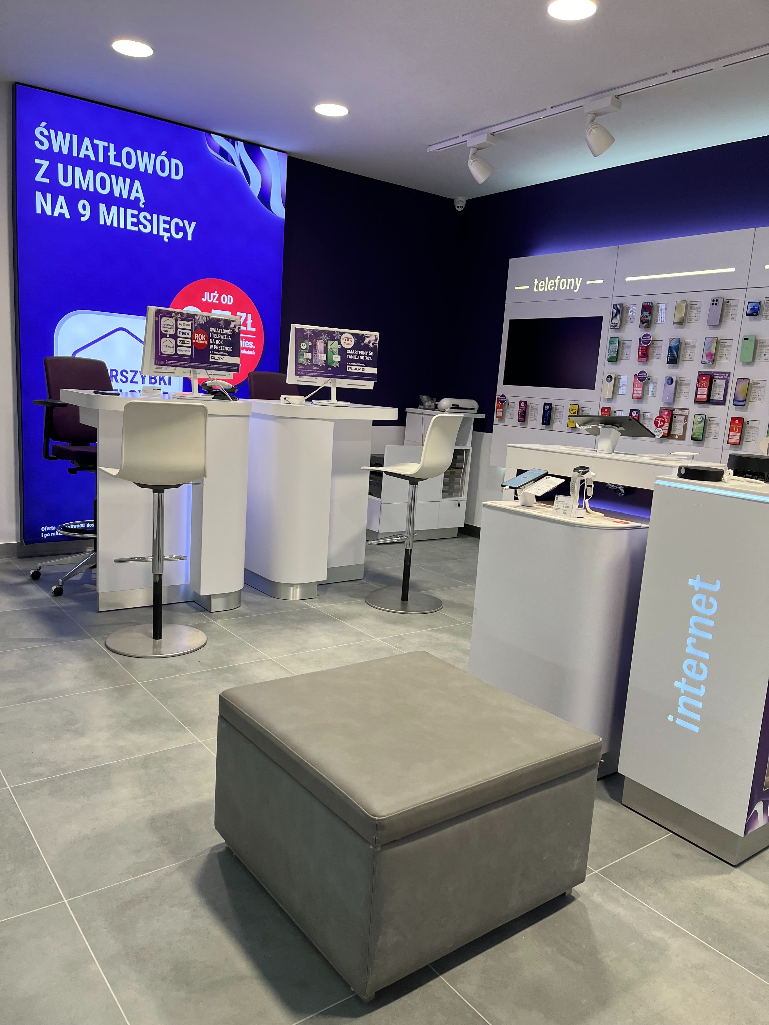 Wnętrze punktu obsługi klienta operatora telekomunikacyjnego w Olsztynie, z widocznymi stanowiskami obsługi, ekspozycją telefonów i dużym ekranem LED prezentującym ofertę światłowodu.