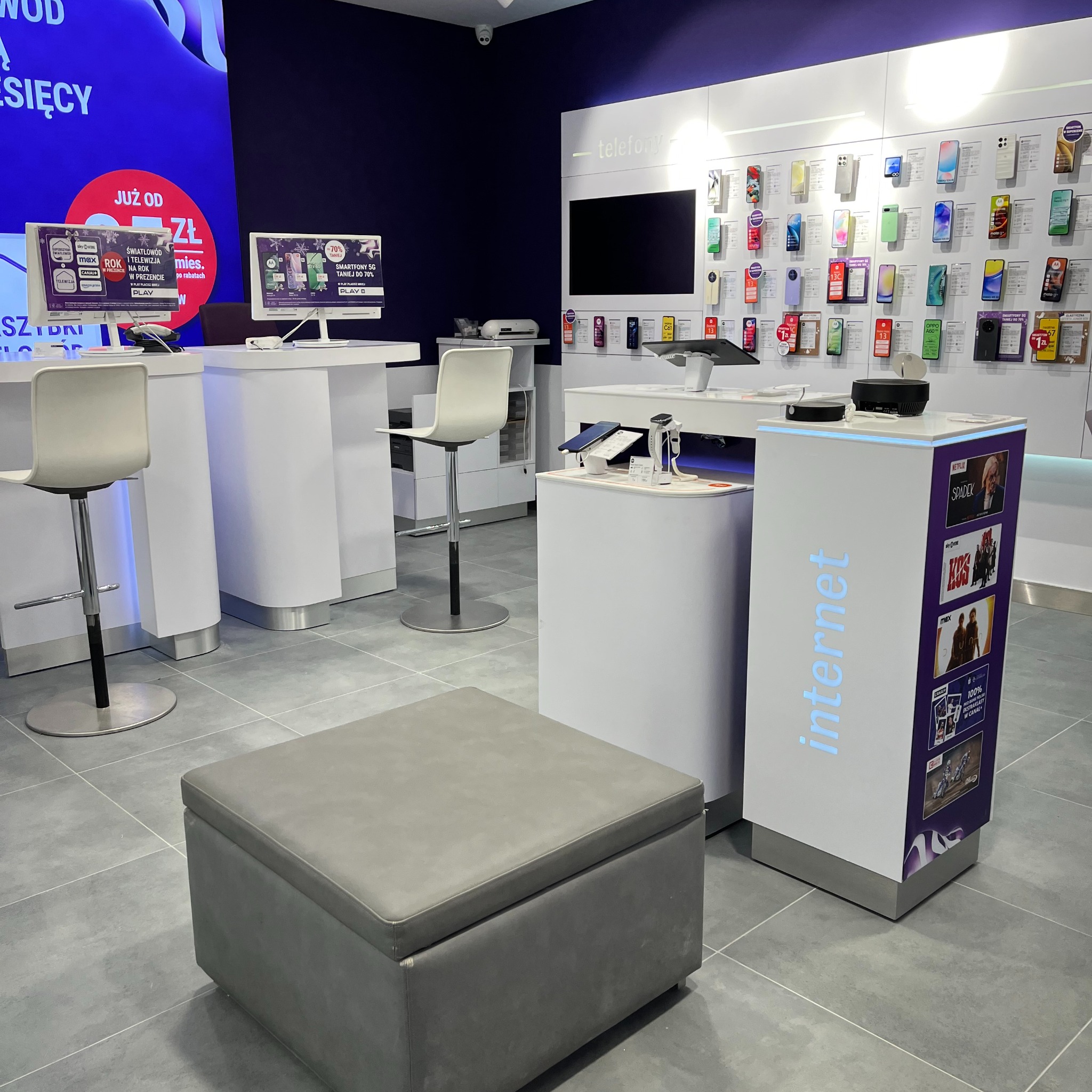 Wnętrze salonu operatora telekomunikacyjnego z ekspozycją telefonów, tabletów i ofert internetowych, widoczne stanowiska obsługi klienta i puf do siedzenia.