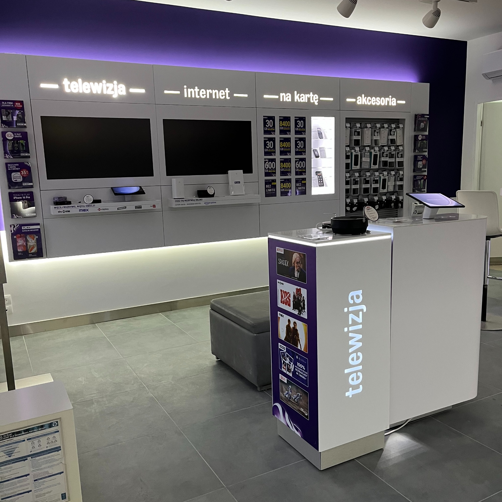 Wnętrze salonu operatora telekomunikacyjnego z ofertą telewizji, internetu i akcesoriów. Na ścianie ekspozycja telewizorów i akcesoriów. Przy ladzie plakaty z ofertami filmów i seriali.